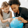 A woman and a young girl look closely at a blue desk globe, with the girl pointing to a specific country.