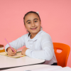 A smiling young girl sits at a school desk with an orange chair, holding a red pencil over her notebook against a pink background.