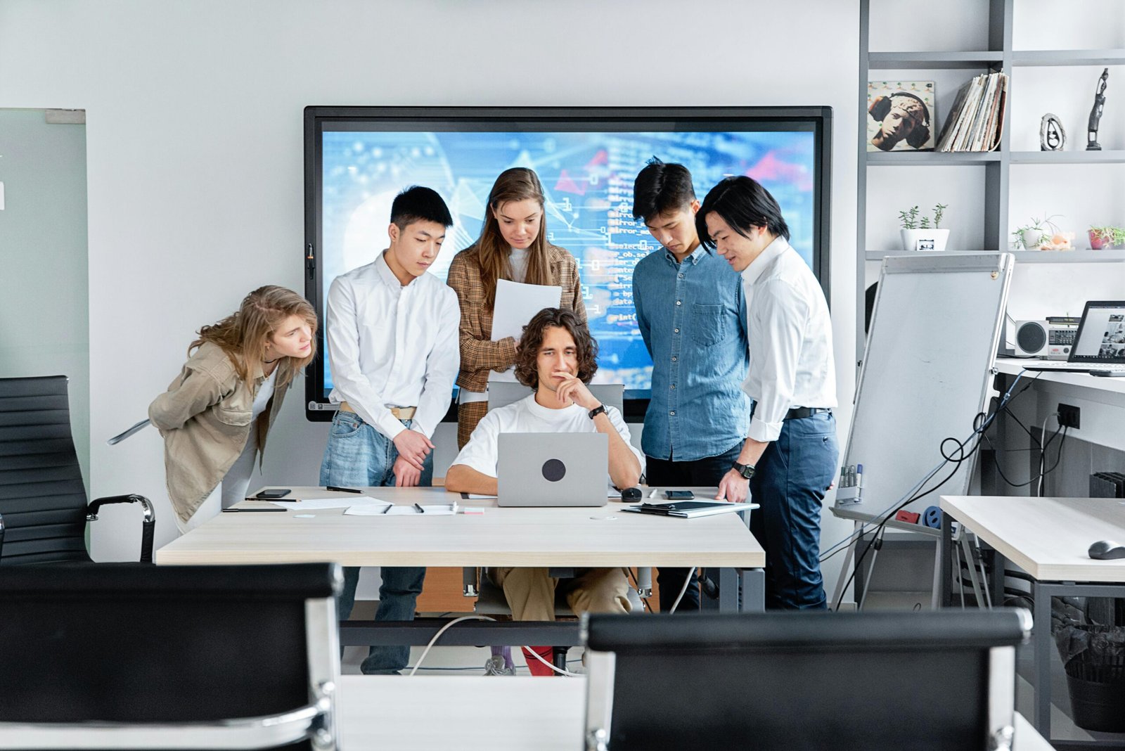 A diverse group of six young professionals gathers around a colleague seated at a desk with a laptop in a bright, modern office.
