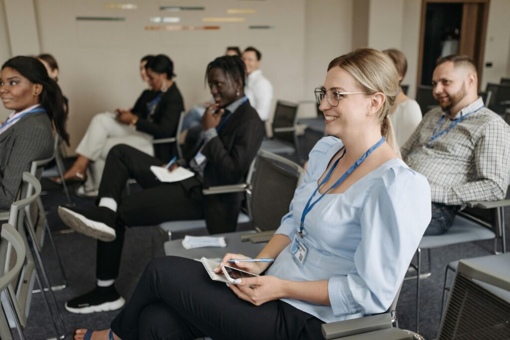A group of diverse young professionals sits in grey chairs at a conference or seminar. In the foreground, a blonde woman wearing glasses and a light blue blouse smiles while holding a notebook and a pen.