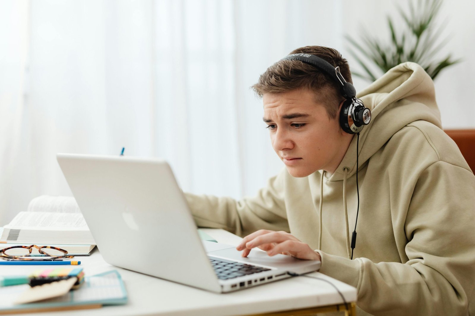 A teenage boy wearing a beige hoodie and headphones leans forward, looking intently at a laptop screen.