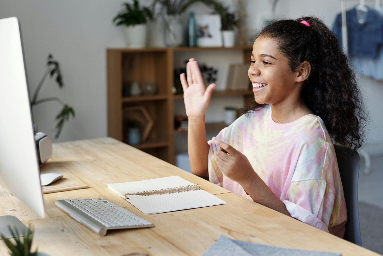 A smiling young girl with curly hair tied in a ponytail waves at a computer monitor while sitting at a light wood desk.
