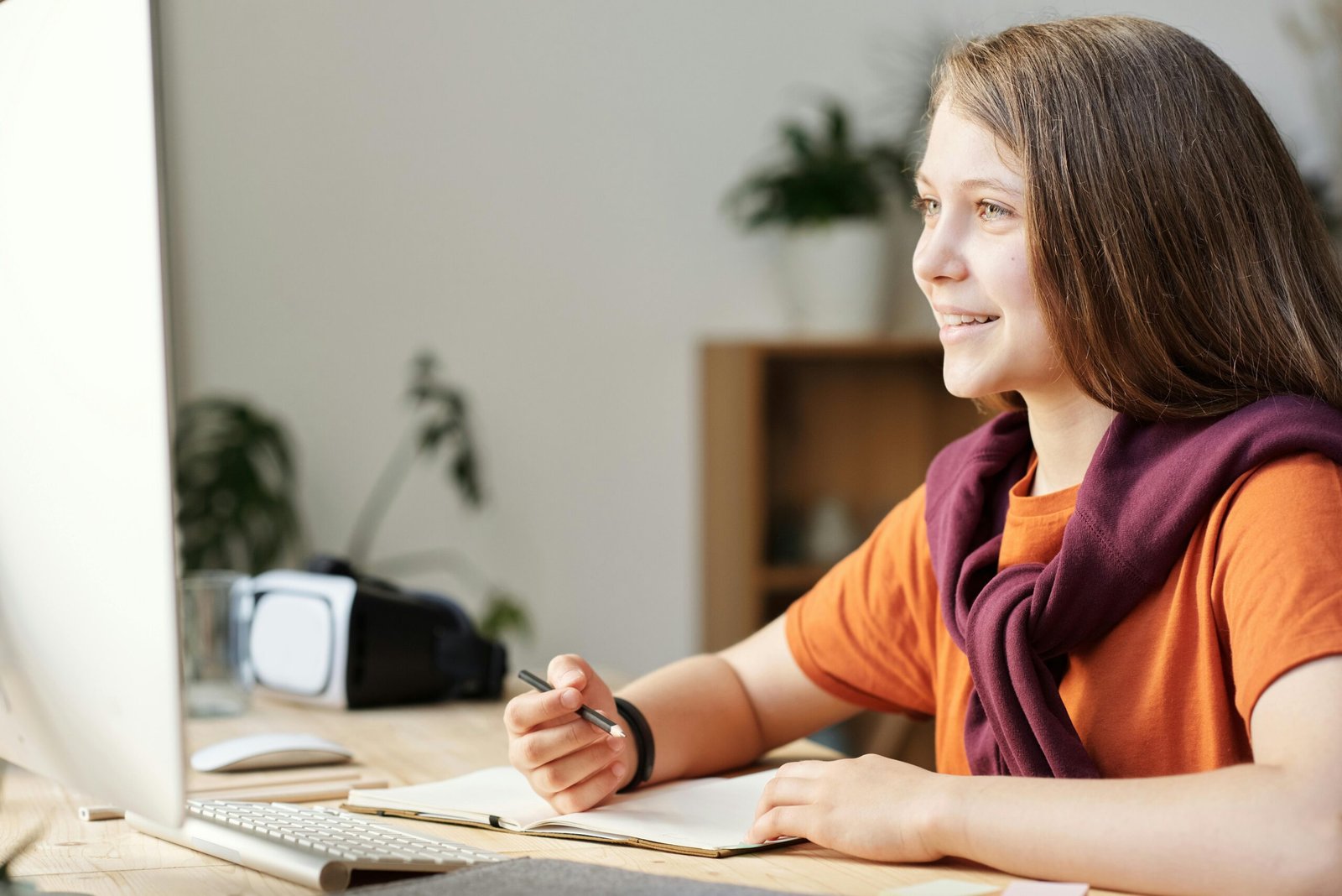 A smiling young girl with long brown hair sits at a wooden desk, looking at a computer screen.