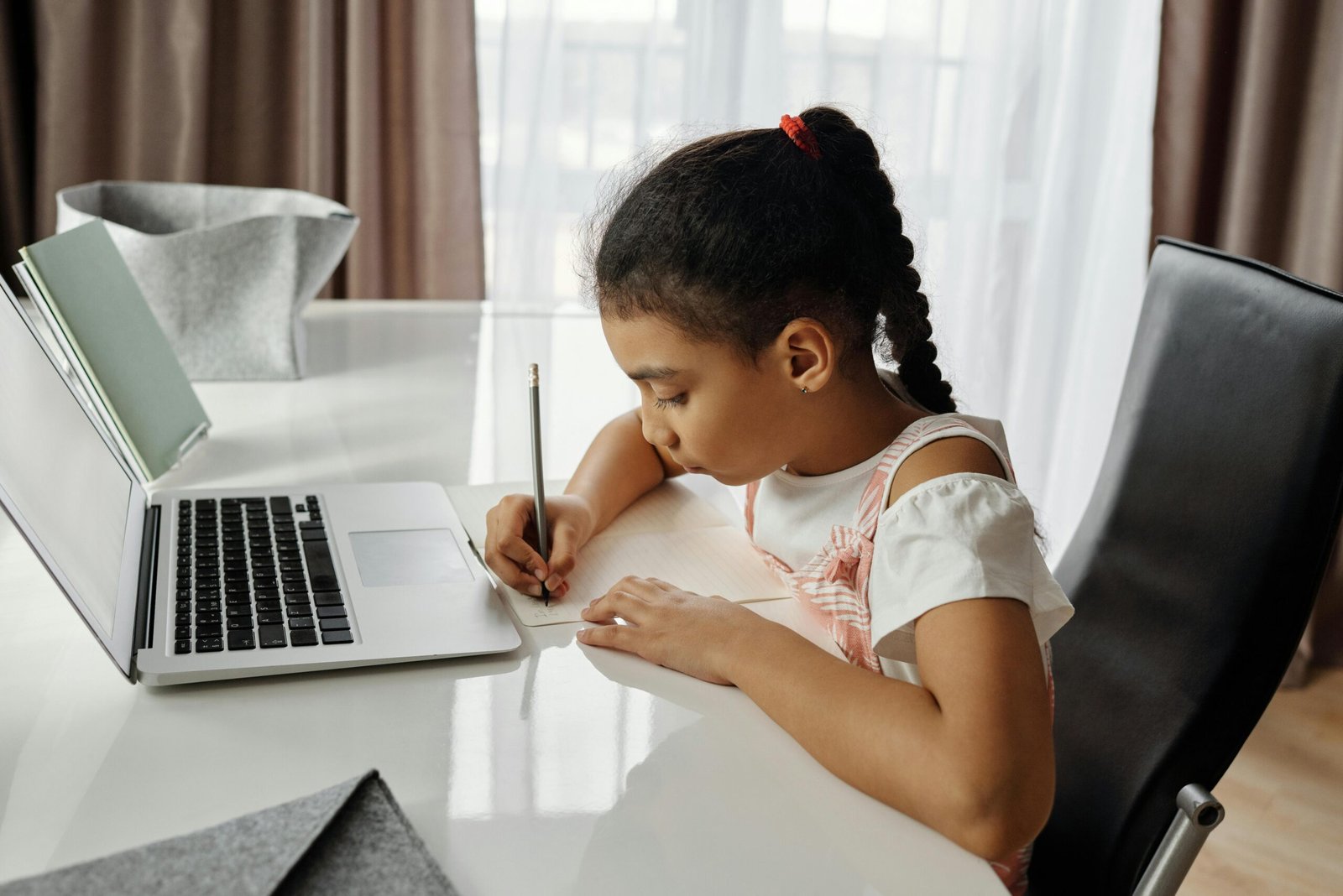 A young girl with braided hair sits at a white desk, writing on a piece of paper with a pencil.