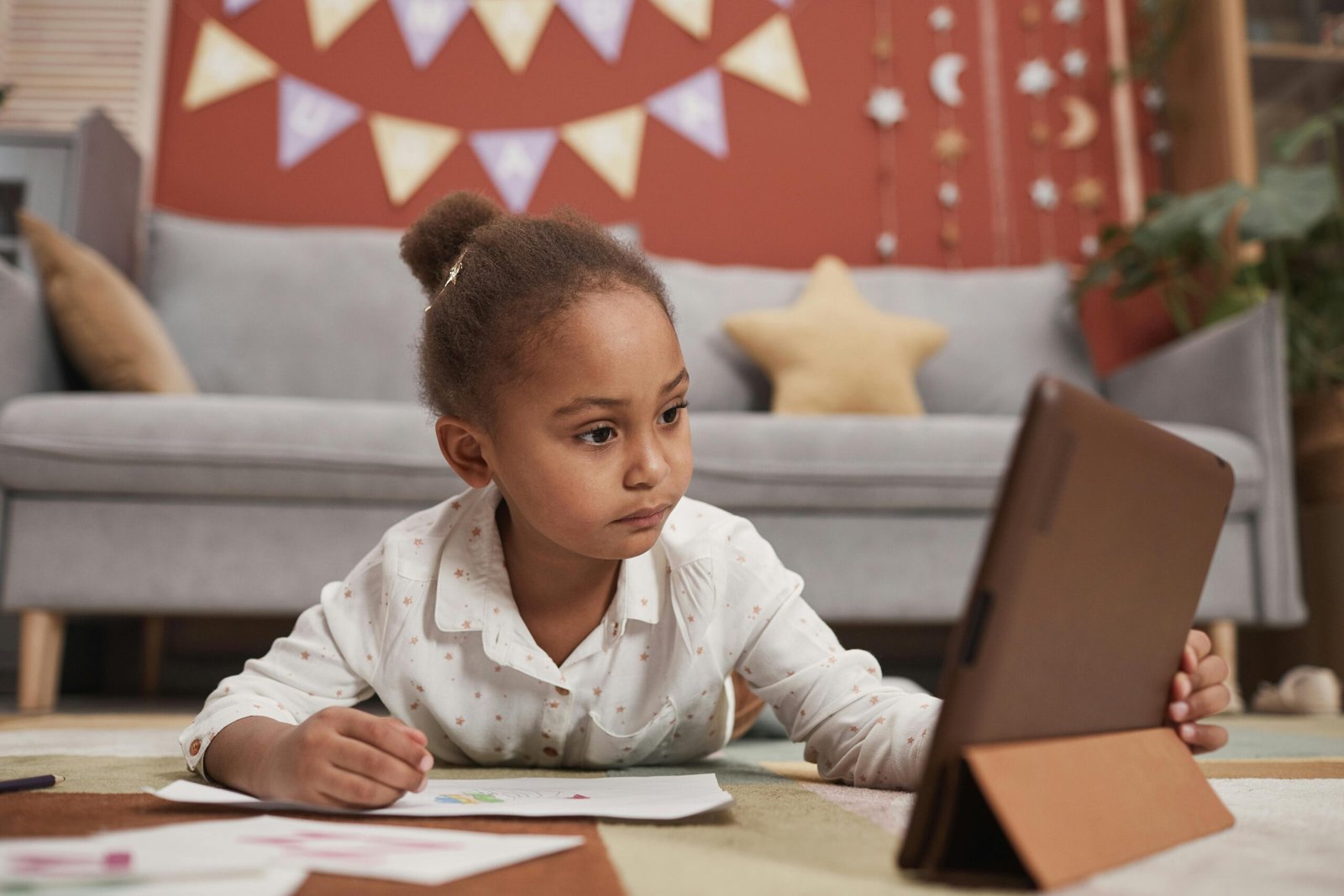 A young girl lies on a colorful rug, looking intently at a tablet held in a brown stand.