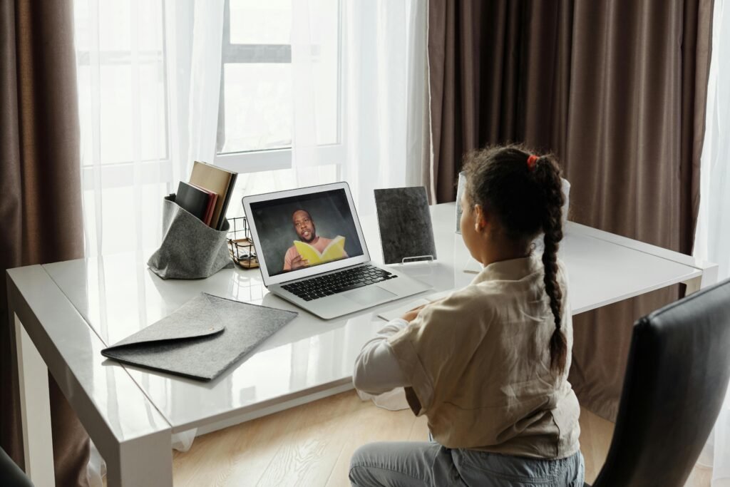 A young girl with a long braid sits at a white desk, facing a laptop showing a man on a video call who is reading from a yellow book.